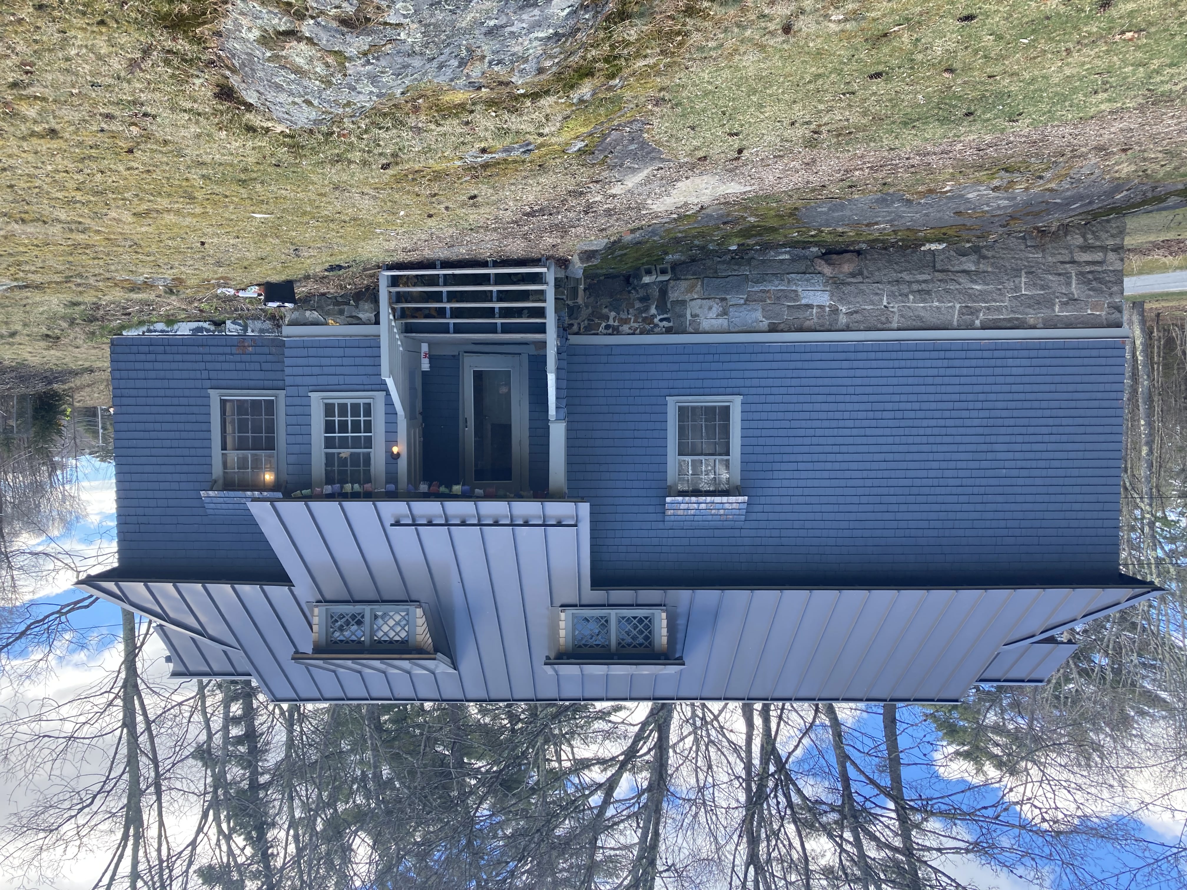 Image of the front of Carriage house. It is a wide blue house with a standing seem metal roof. There is a stone foundation and steps up to the front porch.